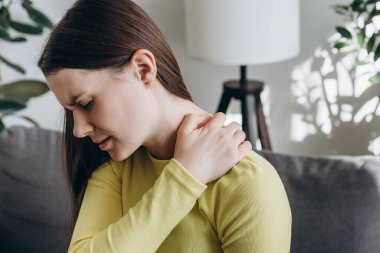 Mutsuz, üzgün, genç bir kadının koltukta oturup omzundaki ağrıdan acı çekmesini ve kendine masaj yapmasını. Boyun ağrısı. Ön kol ağrısı, kas sorunları olan insanlar, sağlık ve tıp konsepti.