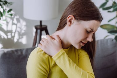 Ağrı, kas sertleşmesi sorunu. Kafkasyalı, üzgün, işten dolayı boynu ağrıyan, masaj yapan, omzunu ovuşturan, evdeki kanepede tek başına oturan genç bir kadın. Sağlık hizmeti kavramı