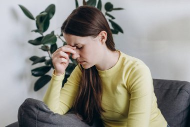 Yorgun ve stressiz genç bayan evdeki kanepede otururken depresyona giriyor. Baş ağrısı, baş ağrısı, baş ağrısı, migren, aşırı çalışma, yorgunluk, bıkkınlık, endişe, endişe, endişe.