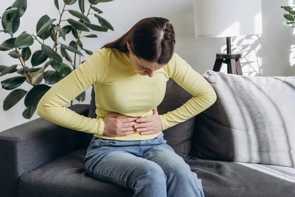 Close up of upset young woman suffering from menstrual pain. Girl with hands squeezing belly having painful stomach ache or period cramps sitting on couch. Abdominal pain and painful periods concept