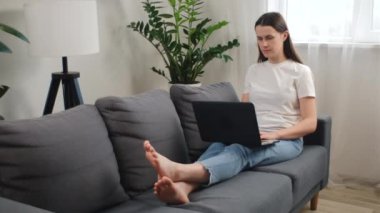 Sakin, serbest çalışan genç bir kadın. Rahat bir şekilde uzanıyor. Klavye kullanıyor ve evdeki rahat koltukta dizüstü bilgisayarda çalışıyor. Girişimci kız oturma odasında işi için çalışıyor. İş çalışma konsepti