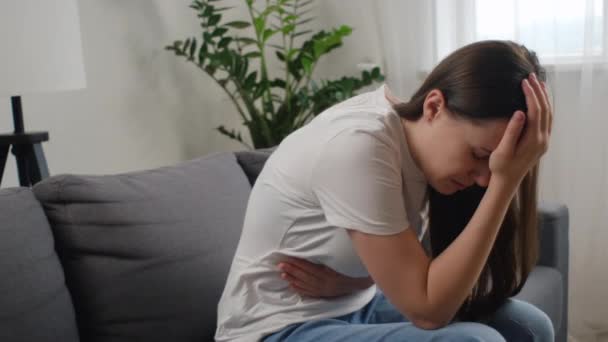 月経痛に苦しんでいる不健康な悲しい若い女性 痛みを伴う胃の痛みを伴う腹を絞った少女またはソファーに座っている期間のクランプ 腹痛と痛みのある期間の概念 — ストック動画