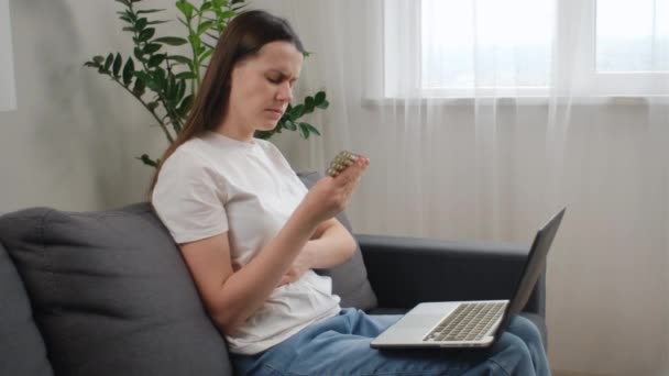 健康とダイエットのコンセプト 自宅でソファーに一人で座っているコンピュータを使用して若い慎重な女性は 錠剤のパックを保持して オンラインビタミン情報に関する処方薬ラベルを読みます — ストック動画