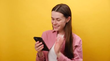 25 'li yaşlarda gülümseyen beyaz bir kadının portresi cep telefonuyla ekranlara bakıyor. Stüdyodaki sarı renkli arka plan duvarının üzerinde tek başına duruyor. Teknoloji, çevrimiçi alışveriş kavramı