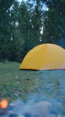Arka plandaki bahar ormanında güzel kamp ateşi ve sarı çadırın dikey videosu. Akşam vakti havada kıvılcımlar uçuşan turist şenliği ateşi. Dışarıdaki yaşam tarzları için ilham verici bir kamp yeri.