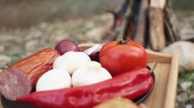 Kırmızı biber, ekmek, domates, soğan, sosis, şampiyonlar, şenlik ateşine hazırlanmak için yemek. Açık hava yemekleri kavramı. Arka plandaki şenlik ateşi ve bahar günü vahşi nehir üzerinde açık yiyecek tabakası.
