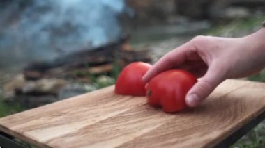 Kadın ellerini mutfak bıçağı kullanarak kapayın. Bahar ormanlarında kamp ateşi ve nehrin yanında tahta kesme tahtasıyla taze kırmızı domates kesin. Sağlıklı beslenme. Dilimlenmiş domates. Kamp kavramı