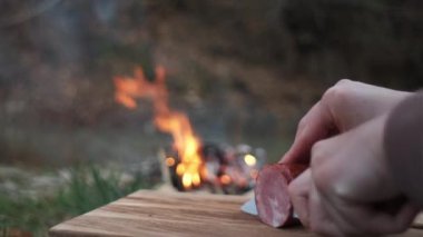Arka plandaki kamp ateşinde ev yapımı et ve sosis doğrayan kadın ellerini ahşap tahtada bıçak kullanarak kapatın. Lezzetli organik domuz eti dilimleyen meçhul genç kadın, köy bahar pikniği.