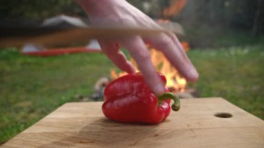Bahar ormanlarında şenlik ateşi ve çadırın yanındaki tahta tahtada taze kırmızı biber kesen genç adama yakın çekim. Yemek pişirme, macera aktif tatiller, seyahat, kamp ortamı ve açık hava yaşam tarzı.