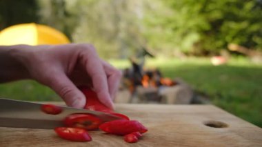 Şenlik ateşinin yanında tahta tahtada taze kırmızı biber ve ormanda çadır keserek erkek ellerini kapat. Kesme tahtasında biber kesmeye hazırlanıyorum. Dışarıda yemek pişirmek. Piknik ve macera konsepti