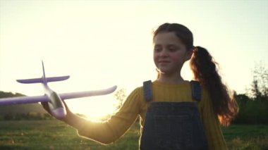 Arka planda oyuncak uçak tutan sevimli, gülümseyen küçük kız çocuğuna yakın çekim. İnanılmaz sıcak günbatımı. Komik çocuk gün doğumunda havacı ya da pilotçuluk oynuyor. Mutlu bir çocukluk. Yavaş çekim