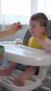 Dikey videoda, sevimli bir çocuğun anne tarafından beslenme, anne tarafından beslenme, sebze yemeği için kaşıkla beslenme, bebek sütten kesme, ilk katı yiyecek. Çocukluk kavramı