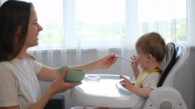 Gülümseyen bir anne kaşıkla sevimli bir bebeği besliyor. Sevgi dolu anne, bebeği kaşıkla meyveli püreyle besler. Yemek zamanı. Küçük çocuk bebek sandalyesinde oturarak yemek yiyor. Anne oğluyla ilgilenir, içeride.