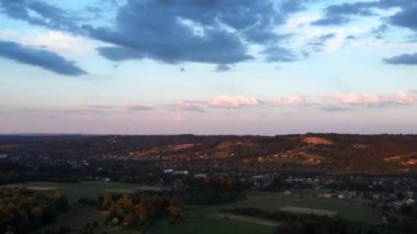 Gün batımında ya da gündoğumunda kırsal kesime yaklaşan orman, ev ve tarım arazilerinin hava aracı görüntüsü. Yaz ya da bahar aylarında kayan tepelerin panoramik manzarası