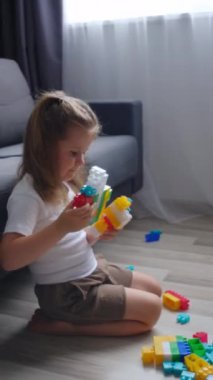 Meraklı küçük kızın renkli plastik tuğlalarla oynadığı dikey video oturma odasındaki kanepenin yanında, sıcak bir zeminde duruyor. Çocuk gelişimi, hafta sonları ev konseptinde boş oyunlar