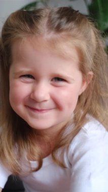 Kameraya bakıp gülümseyen güzel bir kızın dikey portresi. Mutlu şirin çocuk evde rahat bir koltukta oturup eğleniyor, çocukluğun tadını çıkarıyor, keyifli bir ruh hali içinde.