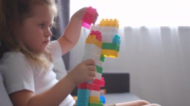 Şirin küçük kız evdeki rahat koltukta oturan renkli inşaatçı oyuncaklarıyla oynuyor. Eğitim oyunu, boş zaman aktiviteleri konsepti. Çocuk anaokulunda inşaatçı küpleriyle oynuyor.