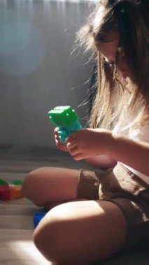 Evdeki sıcak zeminde oturan sevimli küçük kız çocuğunun renkli plastik tuğlalarla oynadığı dikey görüntüler. Çocuk gelişimi, bebek bakıcılığı, hafta sonları ev konseptinde boş oyunlar