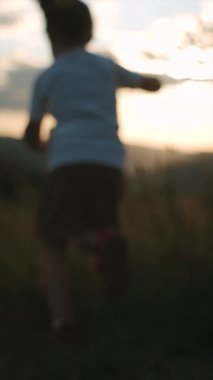 Gün batımında çayırda koşan kız kardeşlerin dikey videosu. Kızlar arka plandaki dağlarda ve gökyüzünde koşarlar. Çocuklar rüya görür. Çocuklar güneşin doğuşunda parkta oynuyorlar. Seçici odak