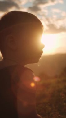 Güzel, sıcak günbatımında arka planda yeşil çimlerde duran sevimli erkek bebeğin dikey portresi inanılmaz dağlardır. Doğadaki küçük çocuk. Parktaki çocuksu rüya konseptin kutlu olsun.