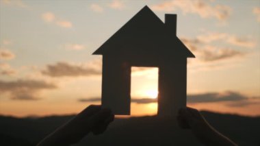 İki kız kardeşin arka planda güzel, sıcak gün batımı ve inanılmaz dağlarda kağıt ev modelini tutarken yakın plan. Yaz doğasında güneş ışığının altında bir yuva. Kendi evini hayal et. Konut ve konut kredisi