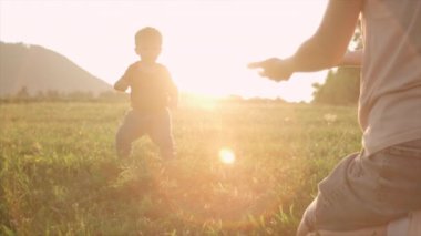 Tatlı bebek oğlu yaz parkında genç annesine gider ve ilk adımları atmayı öğrenir. Çocuk doğada ilk adımları atıyor. Anne oğluna gün batımında arka plandaki dağlarda yürümeyi öğretiyor. Mutlu aile rüya konsepti