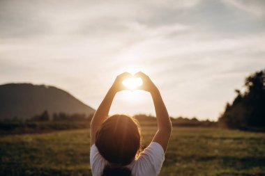 Mutlu küçük bir kız, elleri doğanın arka planında kalp yapıyor. Neşeli çocuk içinde inanılmaz bir günbatımı olan elleri ile kalbini gösteriyor. Tatil, yaz tatili, turizm ve tatil konsepti