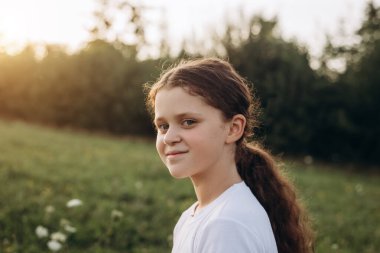 Gülümseyen okul çağındaki genç bir kızın portresi, kameraya mutlu bir şekilde bakmak, sıcak günbatımında güzel doğanın tadını çıkarmak. Açık havada güzel bir çocuk. Çocukluk ve yaz hafta sonu konsepti