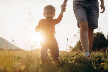 Güzel bebek gün batımında çayırda ilk adımlarını atıyor. Genç anne, arka plandaki dağlarda ileri doğru yürümeyi, evlat gelişimi ve kalkınma kavramını öğrenmesi için çocuğa destek oluyor