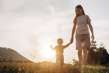 Çocuk doğada ilk adımları atıyor. Tatlı bebek oğlu yaz parkında genç annesine gider ve ilk adımları atmayı öğrenir. Anne oğluna gün batımında arka plandaki dağlarda yürümeyi öğretiyor. Mutlu aile rüya konsepti