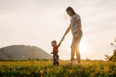 Güzel bebek, gün batımında çimenlerde parkta ilk adımlarını atıyor. Değer veren genç anne, çocuğun arka plandaki dağlarda yürümesini, oğlunun büyümesini ve gelişmesini destekliyor.
