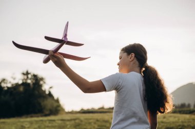 Arka planda oyuncak uçak tutan güzel küçük kız inanılmaz dağlar ve gün batımında gökyüzü. Güneşli bir günde komik çocuk havacı ya da pilotçuluk oynuyor. Mutlu çocukluk kavramı
