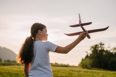 Gün batımında Violet oyuncak uçağıyla oynayan mutlu genç kızın arka görüntüsü. Çocukluk hayalleri. Aile, doğa, özgürlük ve uçak konsepti. Yazın sevimli çocuk pilotu.