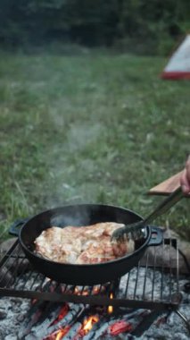 Kamp ateşinde, çadırın yanında ve akşam ormanlarında tavuk göğsü pişirirken çekilmiş dikey bir video. Fırında tavuk göğsünün altın kabuğu, sağlıklı yemek. Açık havada kızarmış tavuk filetosu.