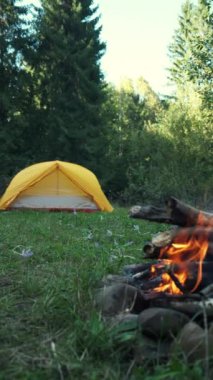 Arka plandaki sonbahar ormanında kamp ateşi ve sarı çadırın dikey görüntüleri. Akşam vakti havada kıvılcımlar uçuşan turist şenliği ateşi. Dışarıdaki yaşam tarzları için ilham verici bir kamp yeri.