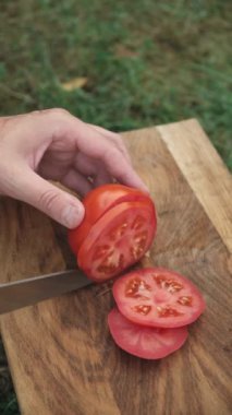 Erkeklerin bıçaklı dikey videosu açık havada tahta tahtada taze kırmızı domates kesiyor. Doğada yemek pişiren bekar bir adam kahvaltı ya da akşam yemeği hazırlıyor.
