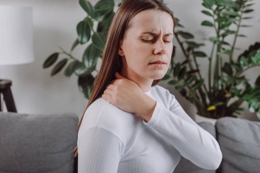 Yorgun, mutsuz, yorgun, boynu ağrıyan, üzgün, sağlıksız beyaz kadının evdeki gri kanepede tek başına masaj yapması. Sağlık kavramı