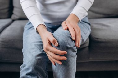 Bacak ağrısı çeken genç bir kadın, kronik hastalıktan dolayı diz ağrısı, el masajı eklem sakatlığı, evdeki kanepede oturan yaralı ya da ağrılı bir kadın. Vücut eklem iltihabı kas sertleşmesi sorunu. Sağlık görevlileri