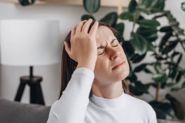 Close up of dizzy young female headache or migraine pain suffering from vertigo or sick, holding head with hands, health problem of brain or inner ear not balance, sitting alone on sofa at home clipart