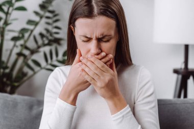 Evde mide bulantısı çeken genç bir kadın, sabah bulantısı olan beyaz bir kadın, oturma odasında kanepede tek başına oturan hasta bir kadın, ağzını kapatıyor.