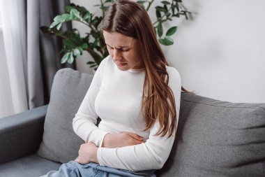 Karnı ağrıyan beyaz hasta kadın karın ağrısı, mide ağrısı, pankreas iltihabı belirtileri. Üzgün kadın koltukta oturup hazımsızlık çekiyor karın şişkinliği sorunu konsepti.