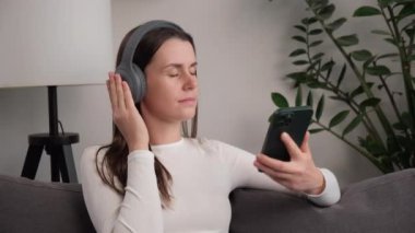 Kulaklık takan, akıllı telefon uygulaması ile müzik dinleyen rahat, sevimli bir kız rahat bir kanepede dinlenen ve cep telefonuyla podcast yapan genç bir kadın.