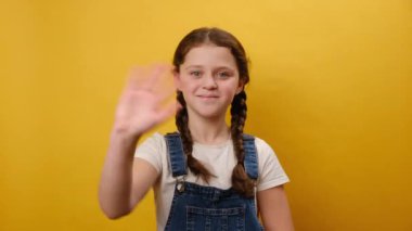 Gülümseyen, el sallayan ve el sallayan küçük kız çocuğunun portresi. Stüdyodaki sarı arka plan duvarında izole edilmiş bir poz veriyor. Çocukluk yaşam tarzı konsepti