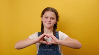 Sevimli gülümseyen küçük kız çocuğunun portresi kameraya bakıyor elleriyle şekil veren kalbi gösteriyor, stüdyodaki sade sarı arka plan duvarında izole bir şekilde poz veriyor. Çocukluk yaşam tarzı konsepti