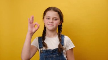 Gülümseyen küçük kız çocuğunun portresi tamam jest yapıyor, kameraya olumlu bakıyor, stüdyodaki sade sarı arka plan duvarının üzerinde izole bir poz veriyor. Çocukluk yaşam tarzı konsepti