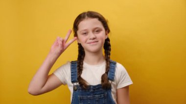 Stüdyodaki sarı arka plan duvarında izole bir şekilde poz vererek zafer işareti gösteren sevimli gülümseyen reşit olmayan kız çocuğunun portresi. Çocukluk yaşam tarzı konsepti
