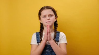 Dua jesti içinde el ele tutuşmuş, bir şey için yalvaran, stüdyodaki sarı arka plan duvarına izole edilmiş güzel bir kızın portresi. Çocukluk kavramı