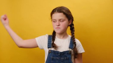 Güçlü, sportif, genç kız işaret parmağıyla pazı kaslarının üzerinde duran portresi stüdyodaki sade sarı arka plan duvarının üzerinde izole bir poz veriyor. Çocukluk yaşam tarzı konsepti
