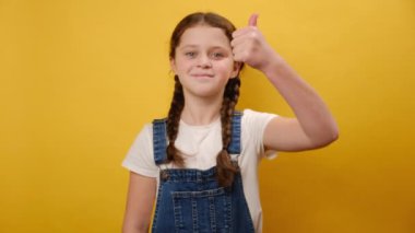 Mutlu gülümseyen küçük kız çocuğunun portresi. Stüdyoda sade sarı arka plan duvarında izole edilmiş bir şekilde duran baş parmağını kaldırma hareketi. Çocukluk yaşam tarzı konsepti