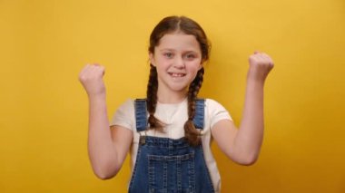 Heyecanlı küçük kız çocuğunun portresi, atölyedeki sade sarı arka plan duvarının üzerinde izole edilmiş bir poz vererek kazananın hareketini yapıyor. Çocukluk yaşam tarzı konsepti